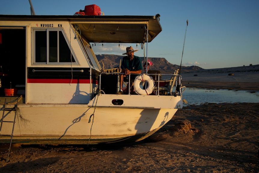Man sits on deck of houseboat at sunset. Boat is in dirt. Small pool of water in background. 