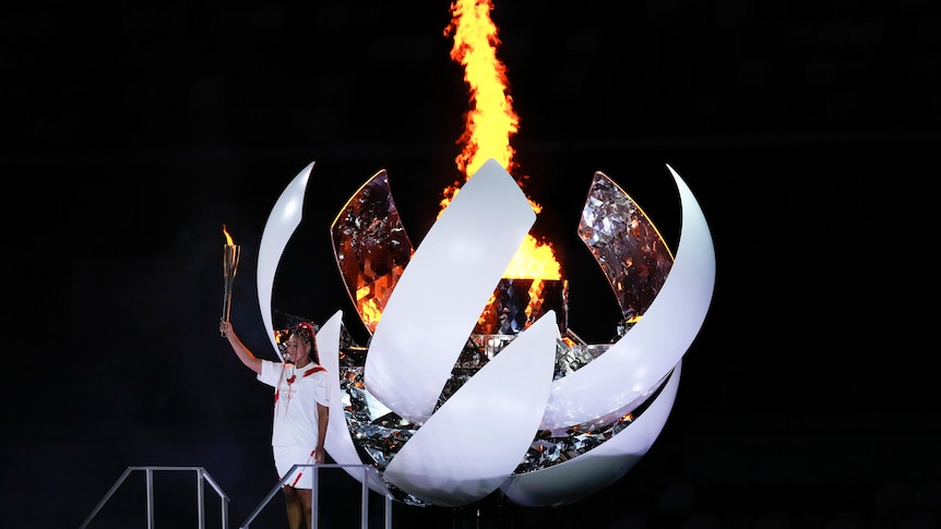 Naomi Osaka stands with a flaming torch aloft next to the burning Olympic cauldron. 