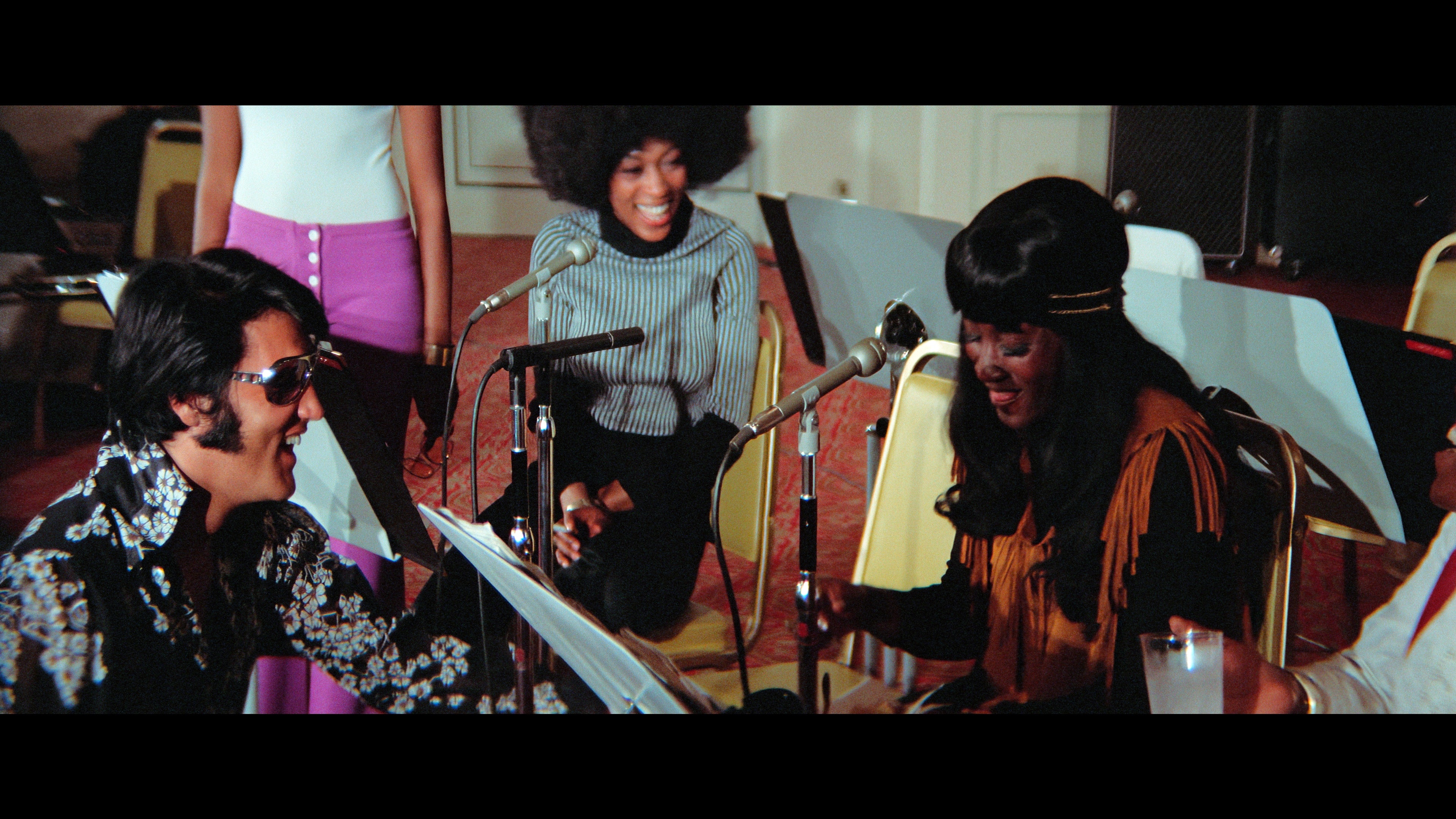 Elvis Presley se ríe en un estudio de grabación, acompañado por cantantes de fondo afroamericanos.