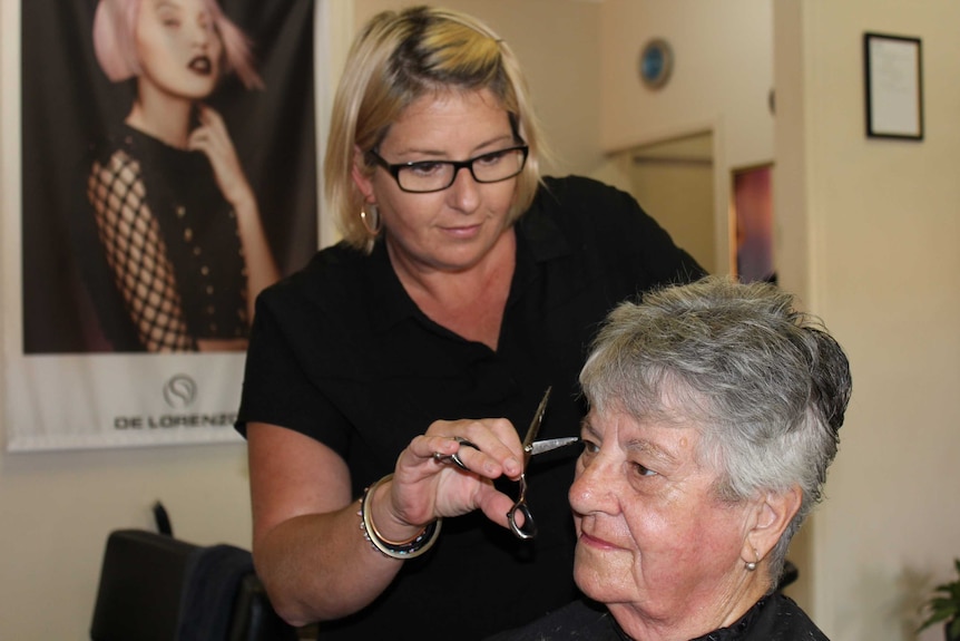 A woman wearing a black dress and spectacles cuts another woman's hair