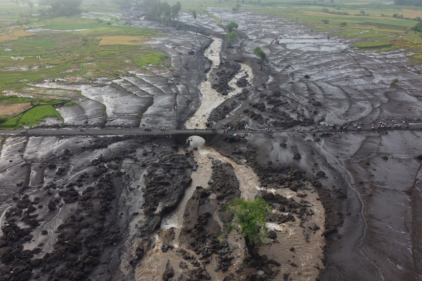 An aerial shot shows dark mud flows from a flooded brown river spreading widely across verdant fields.