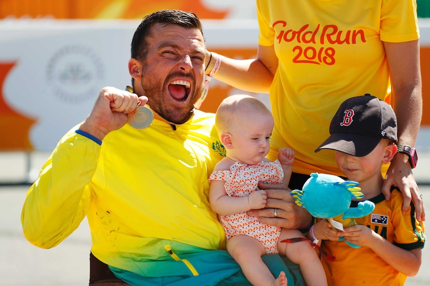 Men in green and gold holds up medal while nursing a baby.