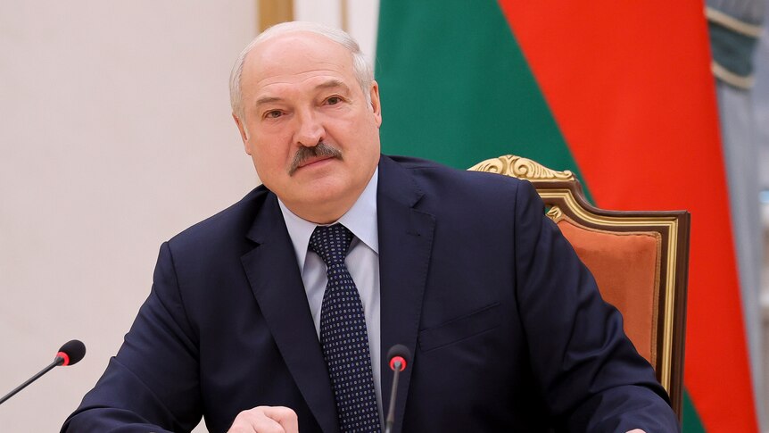 Alexander Lukashenko sits at a desk with microphones. Behind him is the Belarusian flag.
