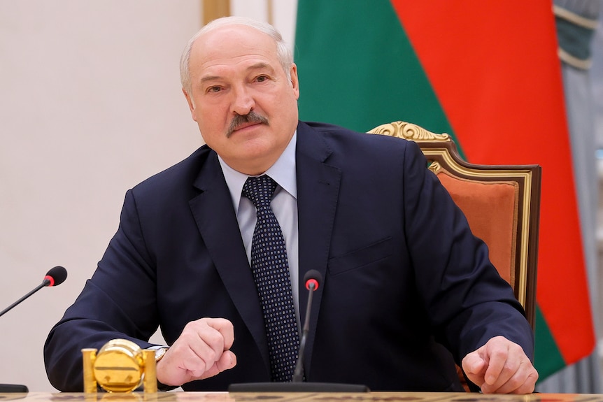 亚历山大·卢卡申科（Alexander Lukashenko）坐在办公桌前，拿着麦克风。 他身后是白俄罗斯国旗。