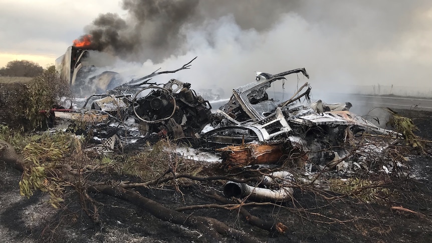 Wreckage of a truck fire on the side of the road. A trailer on fire is in the background.