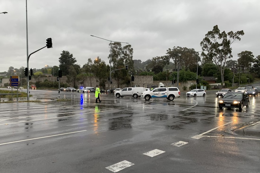 A person in a high-visibility vest directs cars through a rainy major road intersection where traffic lights are not working.