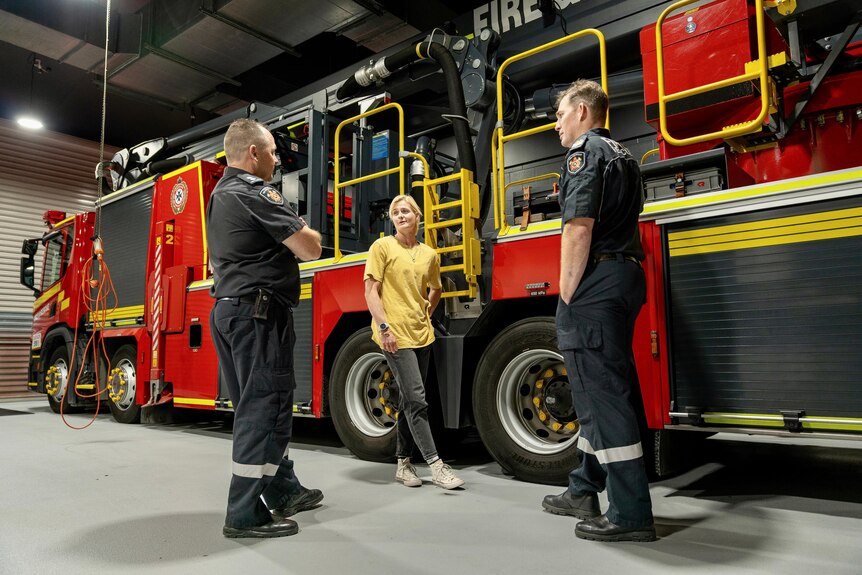 Two uniformed firefighters and a young woman in casual clothes stand in front of a large firetruck.