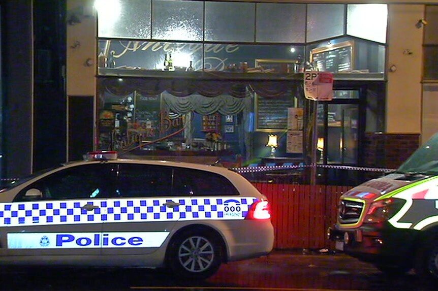 A police vehicle and an ambulance are parked outside a bar with police tape over the front of it at night in drizzling rain.