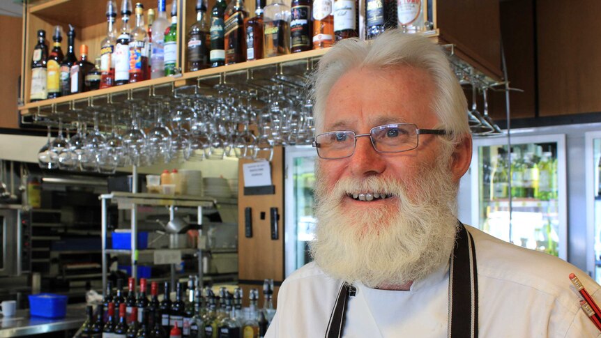 A restaurant owner with white hair and a white beard stands in the venue, wearing a chef's apron.
