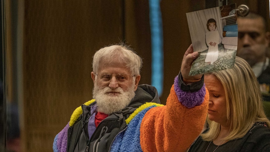 A mamn with white hair wearing a colourful woolly jacket holds a photo of his child in the air.