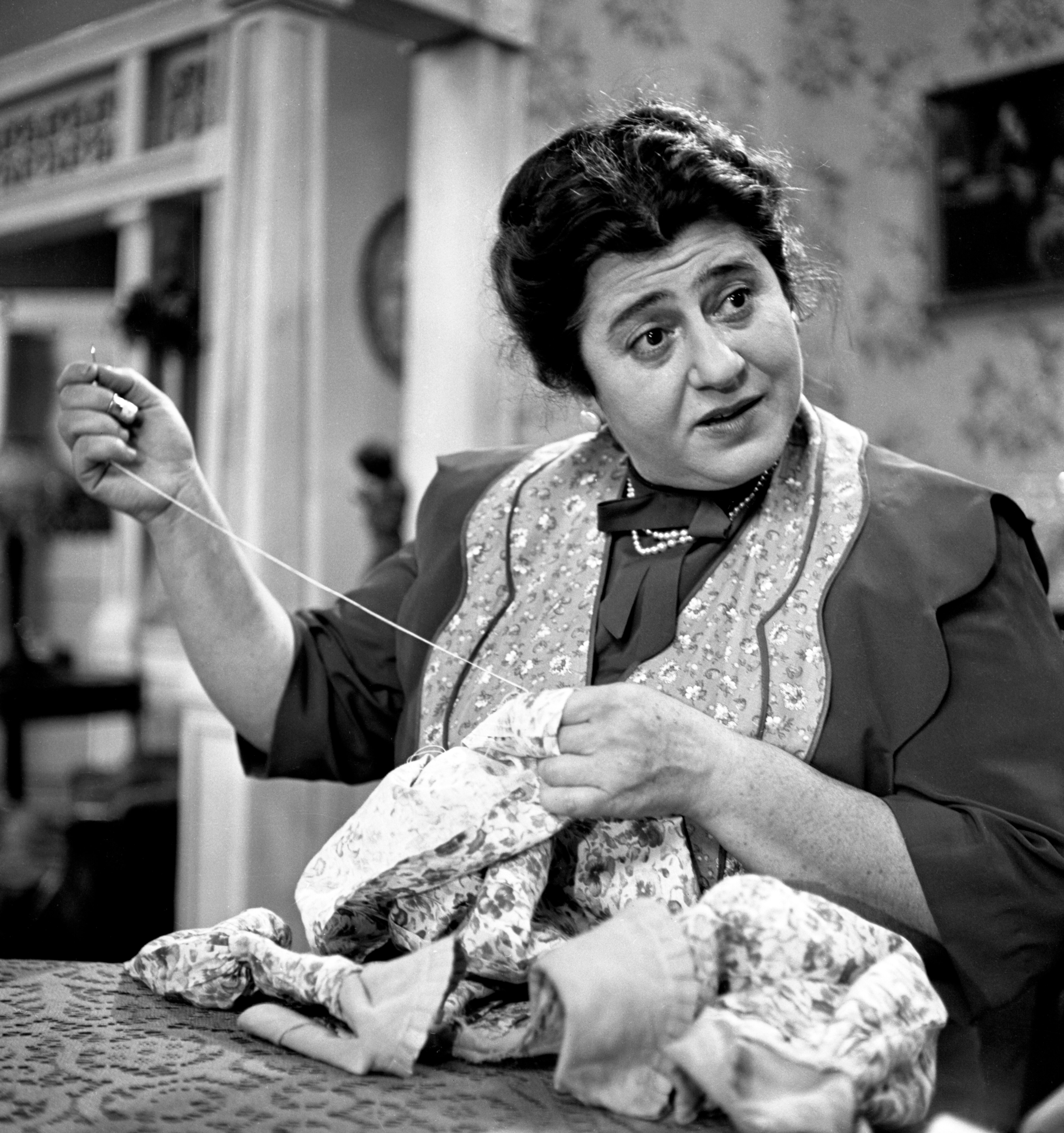 A black and white video still of an older woman looking earnest with her hand on her chest, looking out an open window.