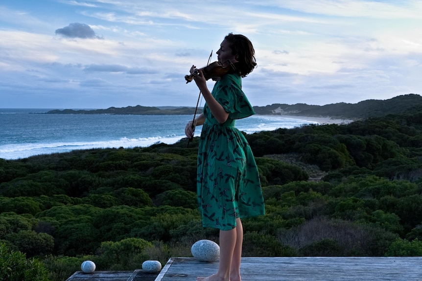 A woman in a green dress plays the violin on a lookout. Behind her is the sea and greenery.