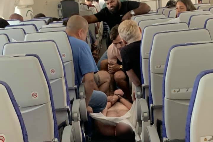 A man lies in an aisle of a plane with his shirt off and people sitting on top of him.