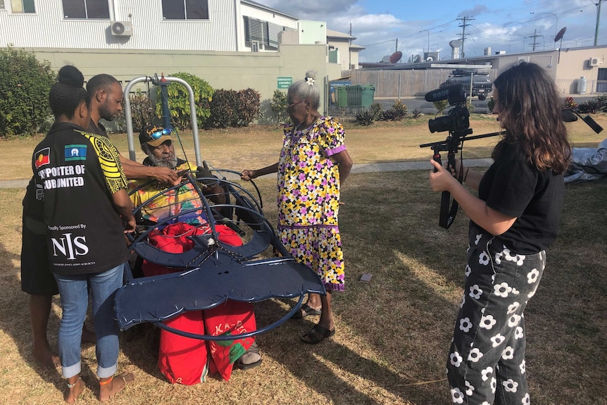 Whitford holding camera filming Thaiday in his wheelchair sculpture structure surrounded by other Indigenous people.