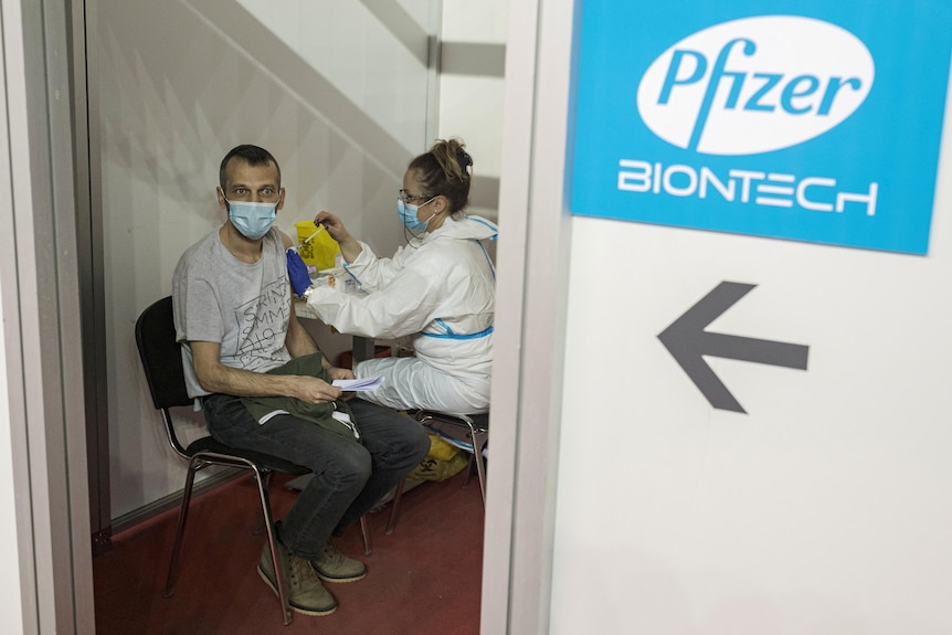 A man is seated as a person in PPE equipment administers a vaccine shot to him.