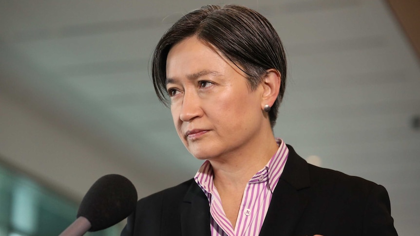 Penny Wong holds a stern expression on her face while people hold microphones and recording equipment in front of her.