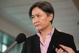 Penny Wong holds a stern expression on her face while people hold microphones and recording equipment in front of her.