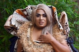 A drag queen dressed in contemporary Fijian cultural attire looks directly forward with a thoughtful expression.