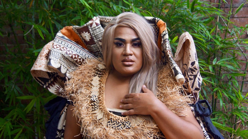A drag queen dressed in contemporary Fijian cultural attire looks directly forward with a thoughtful expression.