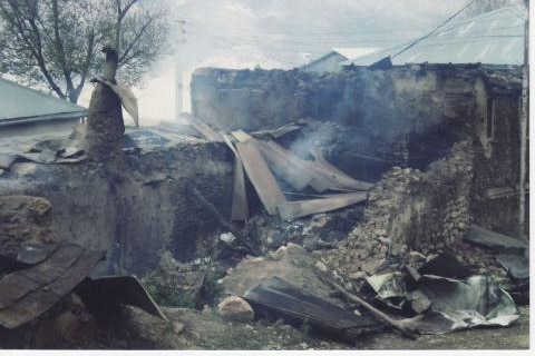 Smoke rises from a house that lays destroyed by a stone fence.