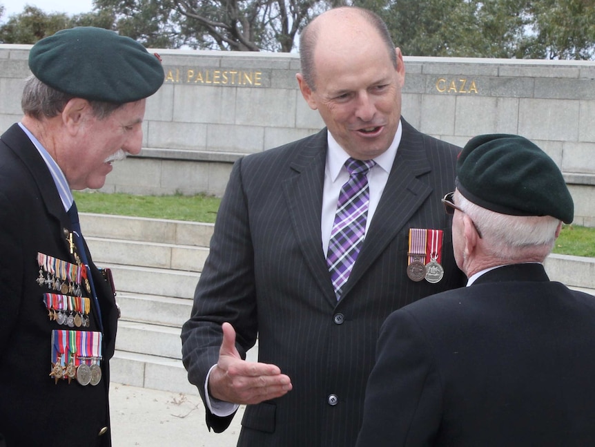 Luke Simpkins stands wearing two war medals talking to two veterans at the State War Memorial.