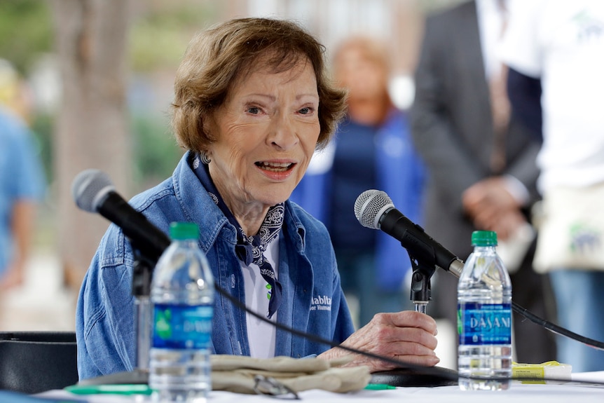 Rosalynn Carter, an older woman wearing a cheerful expression, in front of a microphone.