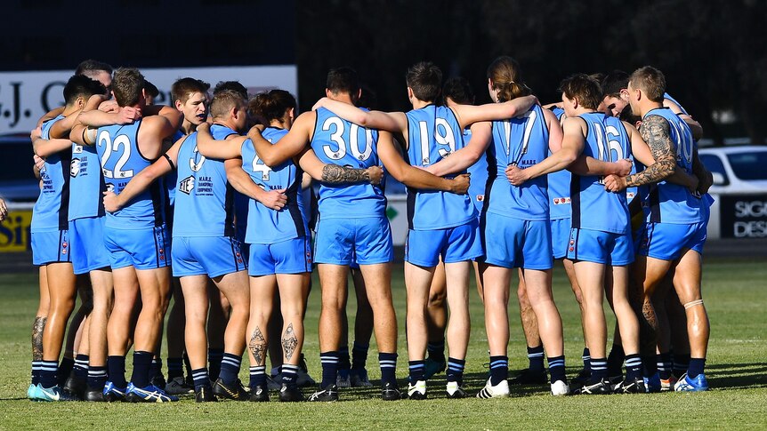 A group of men wearing football uniforms huddle together on oval. 