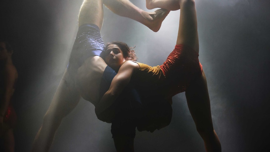 Two acrobats with one foot on the ground and one leg in the air embrace under a white spotlight.
