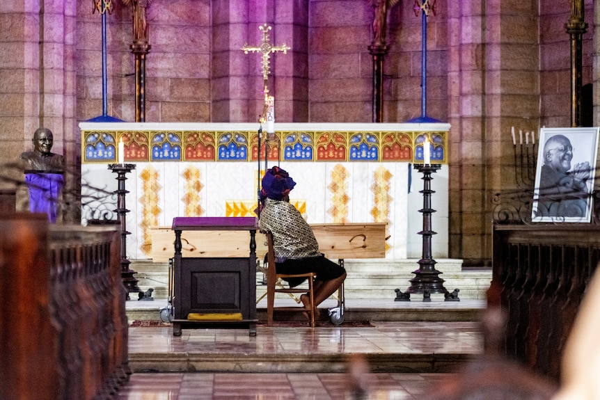 A young woman sits by herself at a funeral in an archaic church