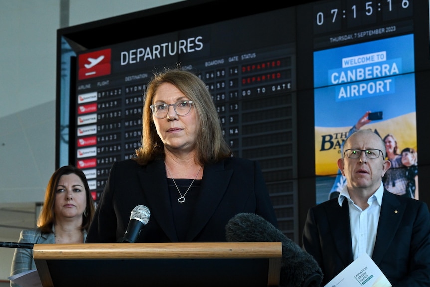 Catherine King in black jacket, with shoulder length blonde hair, stands at lecturn in front of airport signage.