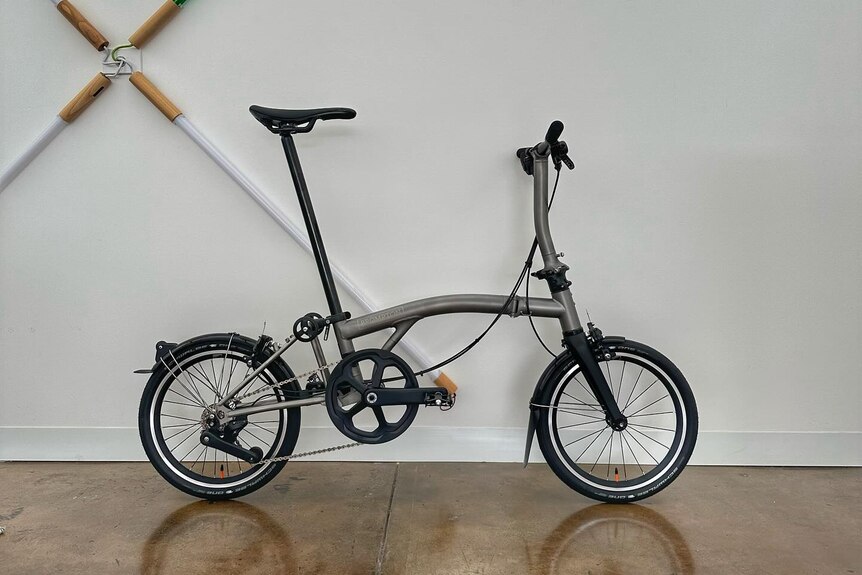 A silver Brompton bicycle stands against a white wall.