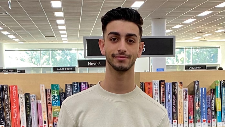 Bassam El Jamal smiles as he poses for a picture in a library wearing a white jumper 