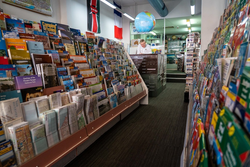 An older man in a white buttoned-up shirt stands at a service desk in a shop space filled with paper maps.