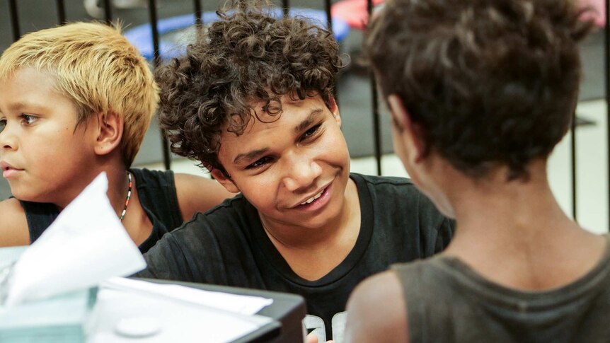 An Aboriginal boy sits facing another boy, smiling as though he is engaged in a conversation.