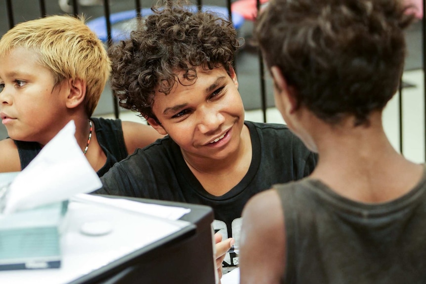 An Aboriginal boy sits facing another boy, smiling as though he is engaged in a conversation.