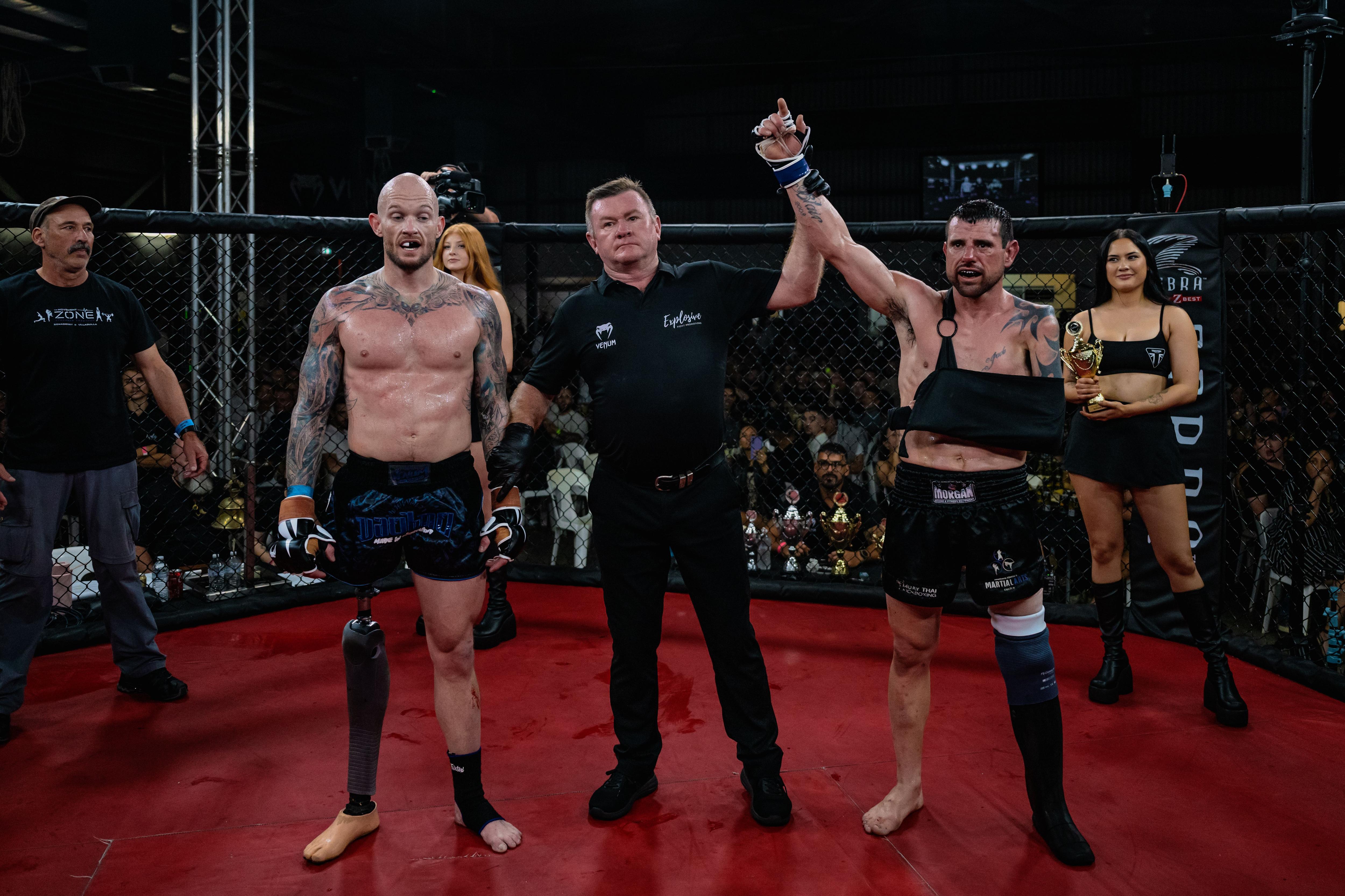 Two fighters, each with a prosthetic leg, stand with a man between them, holding one of the fighter's arms in victory.