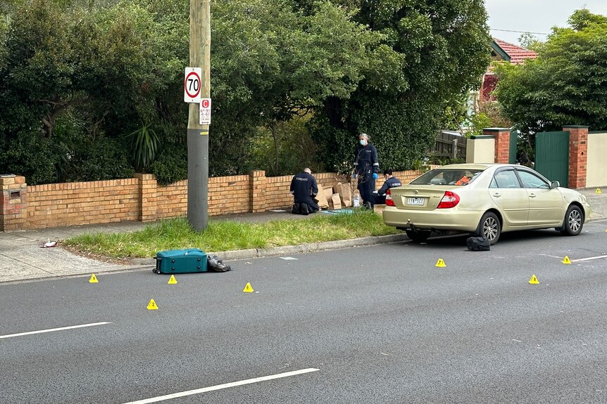 A green suitcase and black plastic bag lie on a road surrounded by yellow police evidence markers with officers on the footpath.