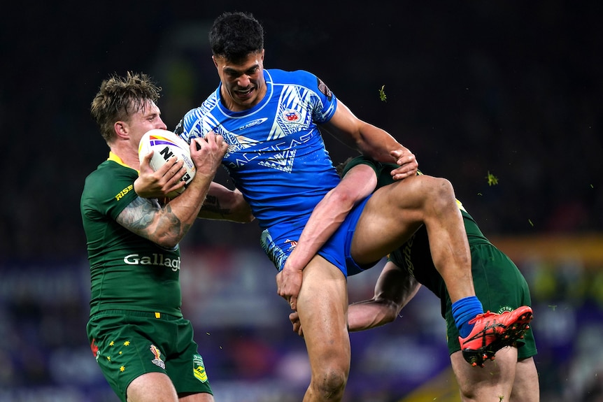 A Samoa male rugby league international carries the ball as he is tackled by two Australian opponents.