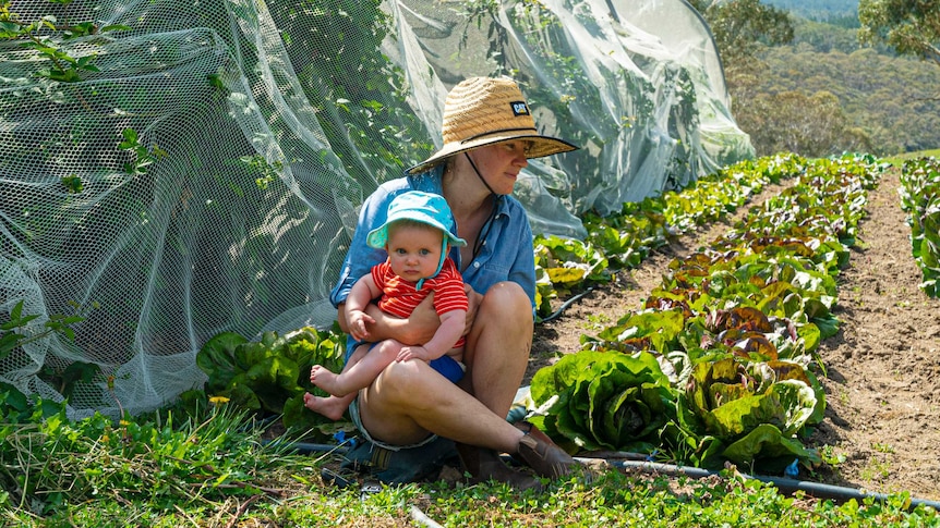 Organic farmer Emily Yarra sitting with her baby beside rows of lettuces growing.