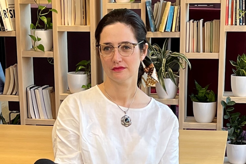 A woman with brown hair wearing glasses sitting in front of an ABC branded microphone. 