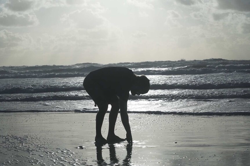 A silhouette of a man bending down to pick up something from the sand.