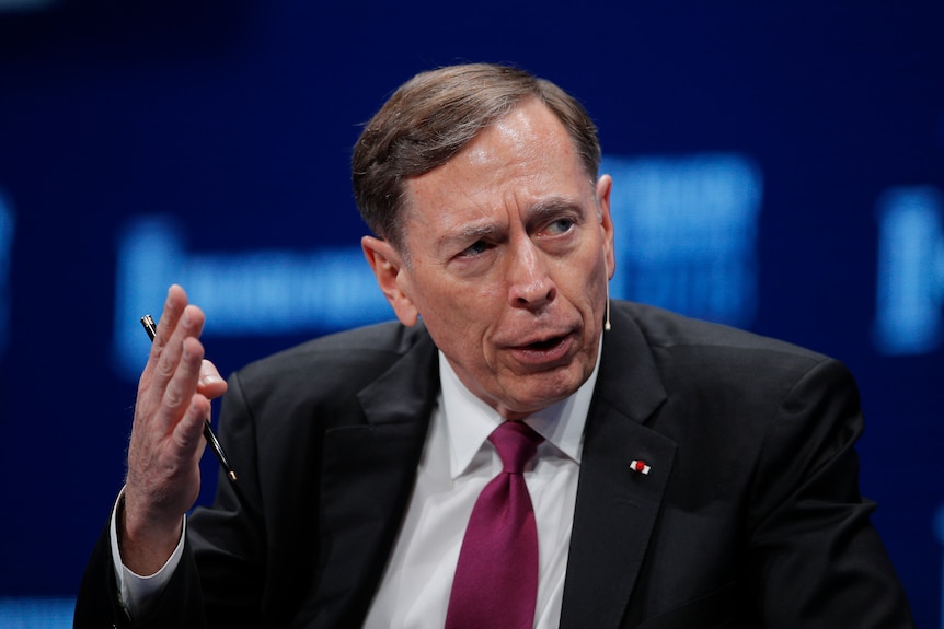 David sits in a suit, speaking and gesticulating with his hand, holding a pen