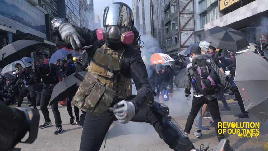 A protester wearing helmet and gas mask gestures towards the camera, others behind use umbrellas to block smoke