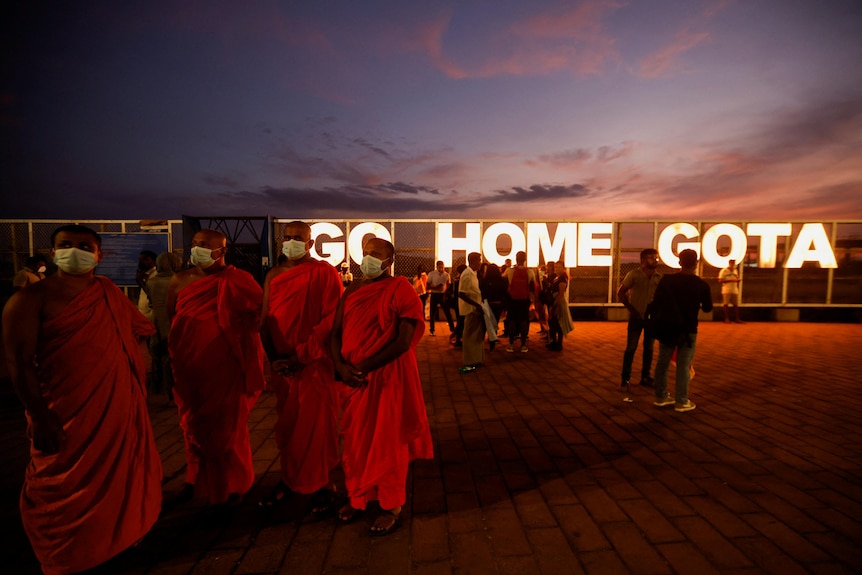 A group of Buddhist monks in orange robes stand next to a lit-up sign reading "Go Home Gota" 