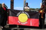 Two women hold an Australian Indigenous flag emblazoned with the words 'don't say sorry, do sorry'.
