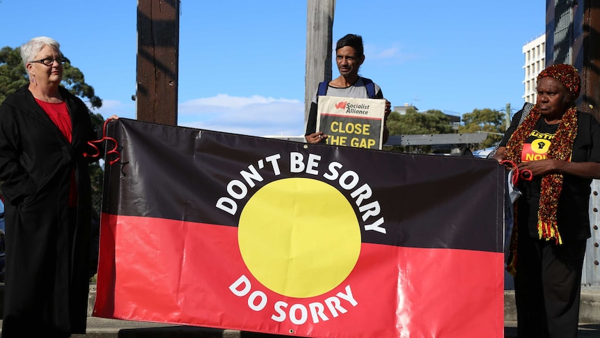 Two women hold an Australian Indigenous flag emblazoned with the words 'don't say sorry, do sorry'.