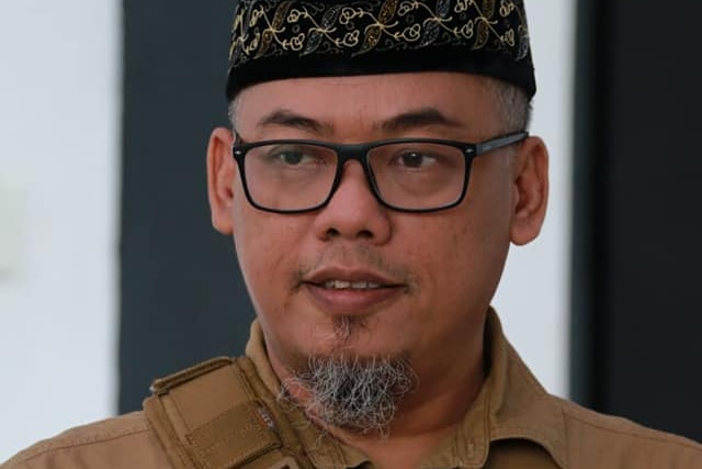 A man in a black hat and glasses, with a thin beard, wearing a khaki shirt smiles at the camera.