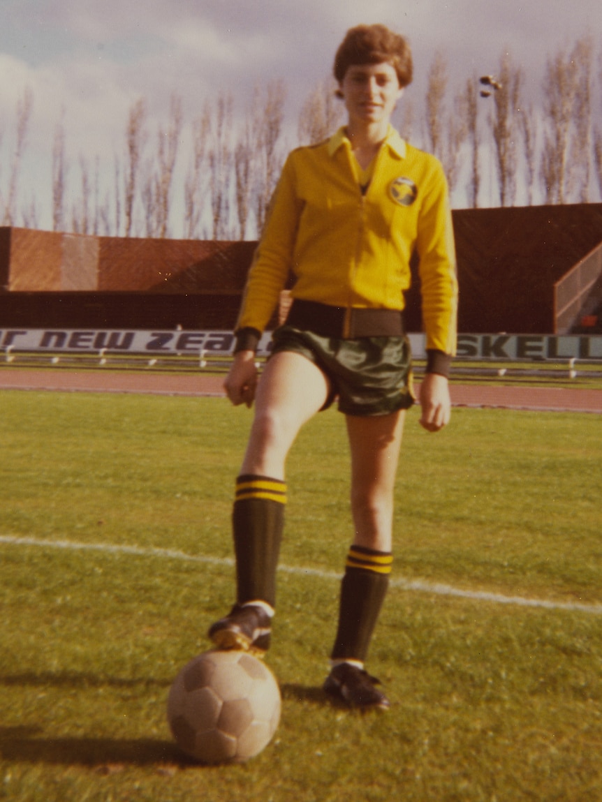 A grainy film photograph of a teenage woman in gold and green with her foot on a soccer ball 