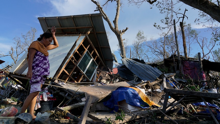 A woman puts her left wrist on her forehead as she moves amongst the wreckage of a home, which has been totally flattened.