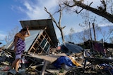 A woman puts her left wrist on her forehead as she moves amongst the wreckage of a home, which has been totally flattened.